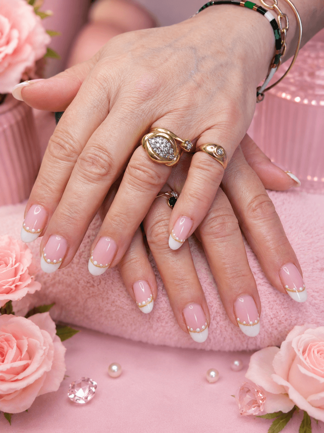 Mains avec manucure française perlée, bagues dorées et décor floral rose élégant.