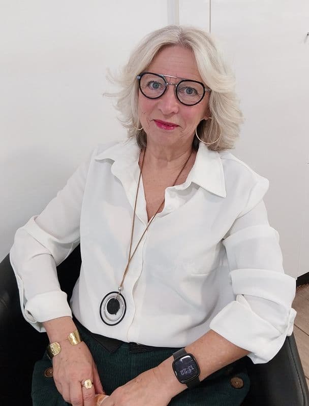 Femme mûre aux cheveux blonds, lunettes noires et chemise blanche, portant des bijoux élégants.