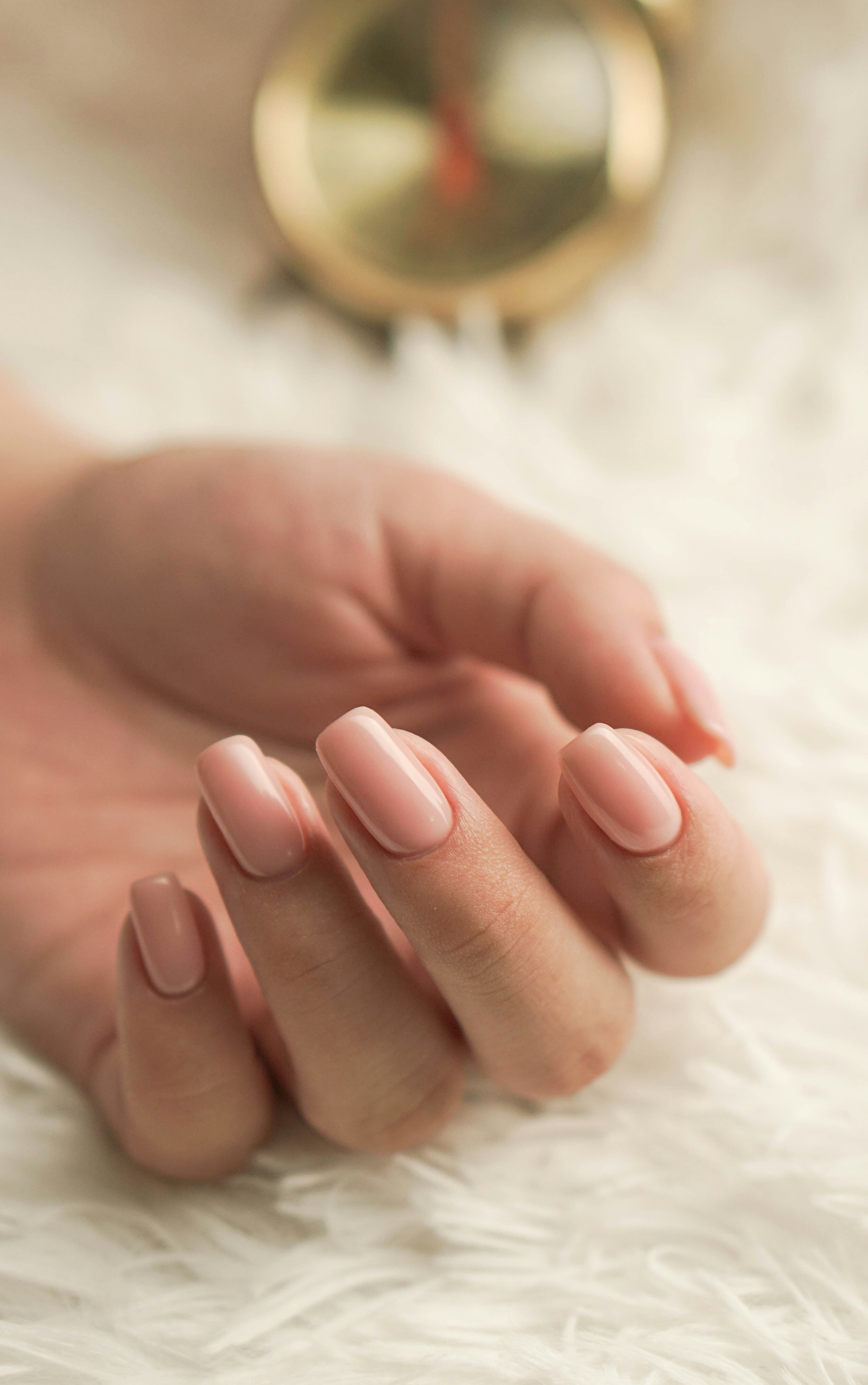 Main avec manucure rose nude brillant sur une surface blanche duveteuse, arrière-plan doré flou.